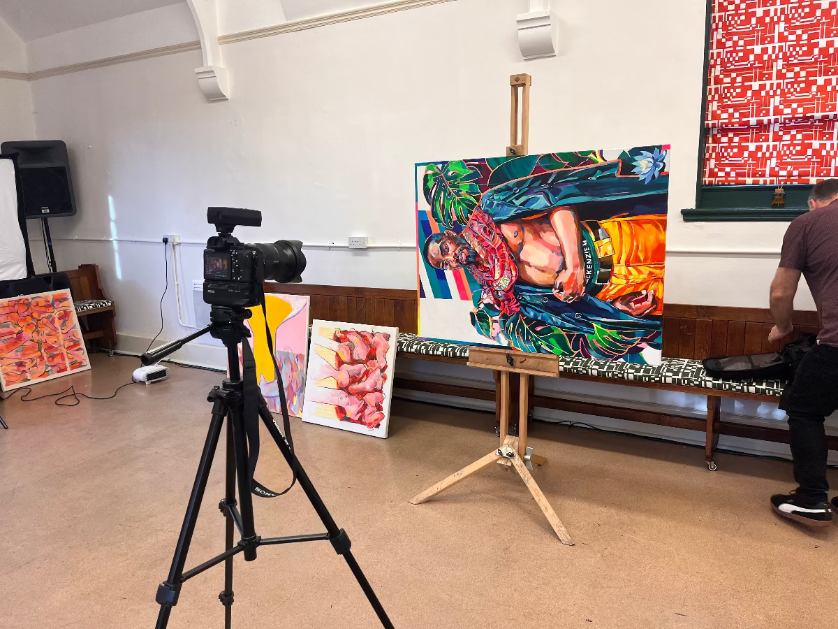 Art studio with a camera on a tripod, easel with colorful painting, and person in the background.