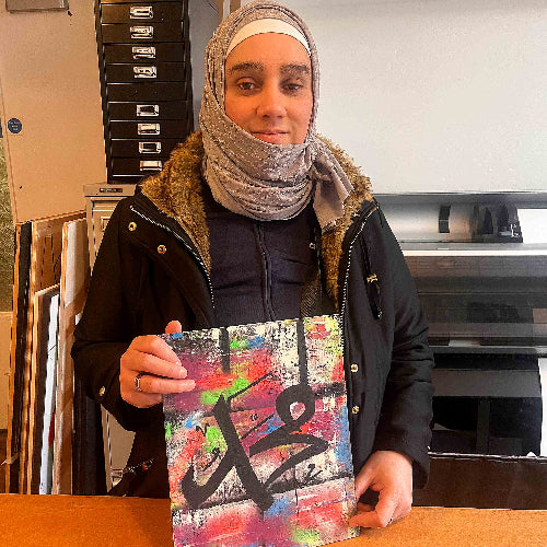 Person holding a colorful artwork with Arabic calligraphy in an indoor setting.